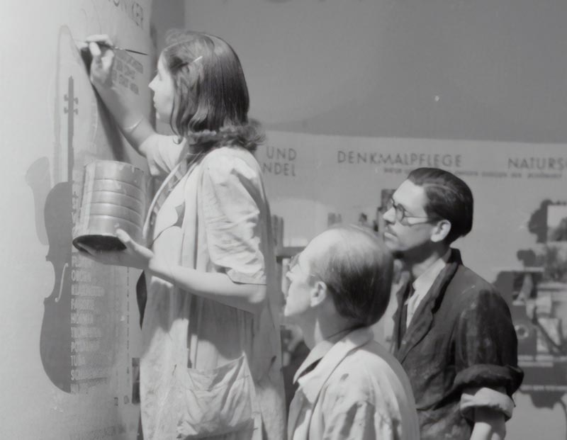 Una mujer está de pie en un escalón y escribe o pinta en una pared mientras sostiene un recipiente; dos hombres están cerca observando su trabajo. En la pared hay texto y diagramas de fondo.