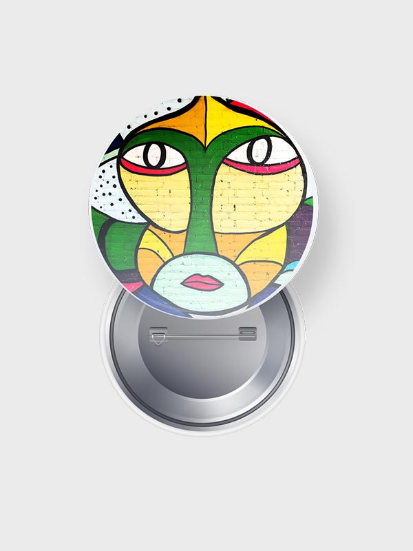 Insignia de botón redondo con un colorido arte callejero abstracto de una cara con grandes ojos verdes, labios rojos y formas geométricas, sobre un fondo liso de color gris claro.