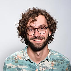 Un hombre sonriente con el pelo castaño rizado, barba y gafas redondas lleva una camisa estampada de colores y está de pie delante de un fondo liso de color gris claro.