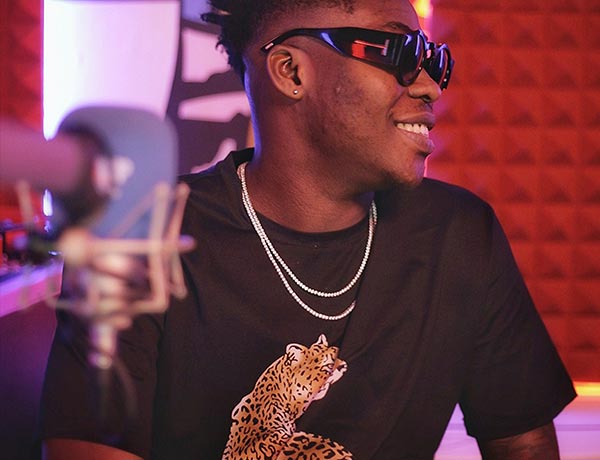 Un hombre sonriente con gafas de sol oscuras, camiseta negra con un gráfico de leopardo y cadenas plateadas está sentado en un estudio de grabación con paneles acústicos rojos y un micrófono en primer plano.