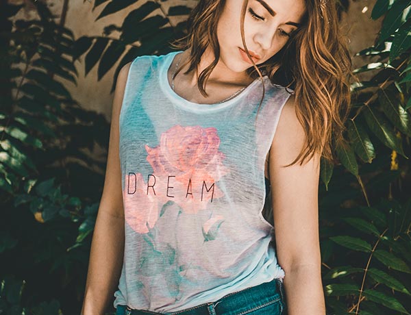 Una mujer de pelo castaño ondulado mira hacia abajo mientras está de pie entre hojas verdes, llevando una camiseta sin mangas con un gran gráfico de una rosa y la palabra DREAM impresa en ella.