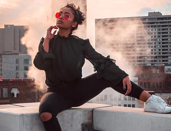 Una persona con gafas de sol rojas, un top negro con volantes y unos vaqueros negros rotos posa con confianza en la cornisa de un tejado con una pierna levantada, rodeada de vapor, con un telón de fondo de edificios de la ciudad al atardecer.