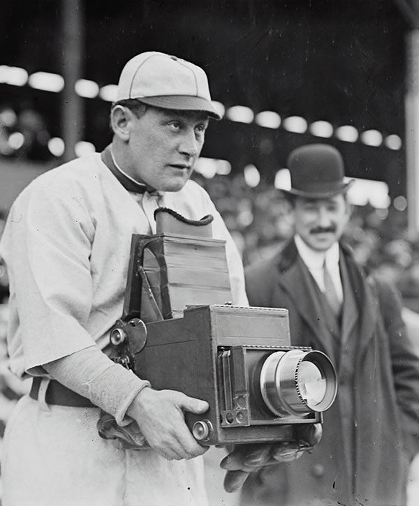 Una foto de época muestra a un jugador de béisbol en uniforme sosteniendo una gran cámara de caja antigua, con un hombre con traje y bombín de pie detrás de él en un estadio lleno de gente.
