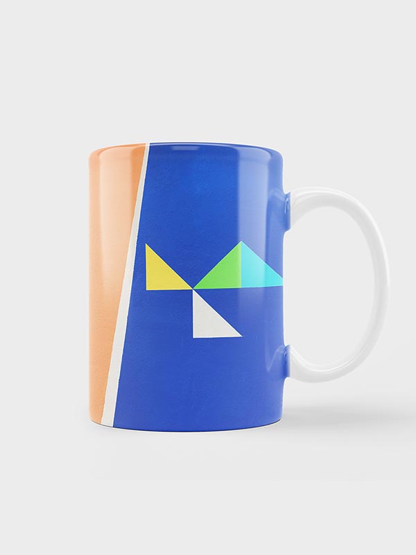 Taza de cerámica con asa blanca y diseño geométrico con formas naranjas, blancas, azules, amarillas, verdes y azul claro sobre fondo azul.