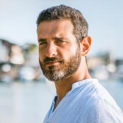 Un hombre con pelo corto, rizado y oscuro y barba está de pie al aire libre y lleva una camisa azul claro. El fondo está difuminado y muestra agua, barcos y edificios bajo un cielo despejado.