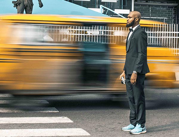 Un hombre con traje negro y zapatillas azules se encuentra en un paso de peatones mientras un vehículo amarillo pasa a toda velocidad, creando un efecto de movimiento borroso. Lleva algo en la mano derecha y mira al frente con calma.
