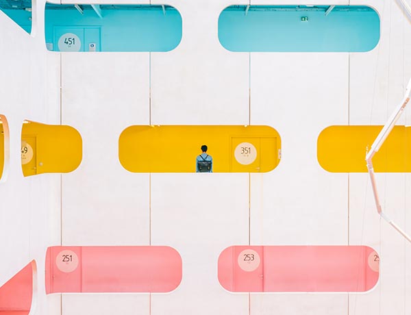 Una persona se encuentra en un pasillo amarillo de un edificio moderno con coloridas ventanas de forma ovalada y puertas azules, rosas y amarillas en diferentes plantas, cada una etiquetada con un número de habitación de tres dígitos.