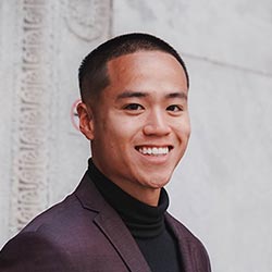 Un joven de pelo corto sonríe a la cámara, vestido con una americana oscura y cuello alto negro, de pie frente a un muro de piedra clara con tallas decorativas.