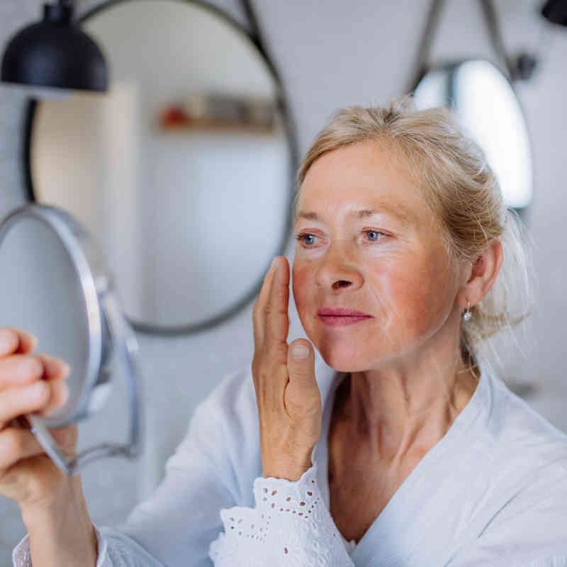 Una mujer mayor rubia con bata blanca se aplica Albura rosa crema nutritiva con vitamina A 200ml en la mejilla mientras se mira en un pequeño espejo en un baño luminoso y moderno.