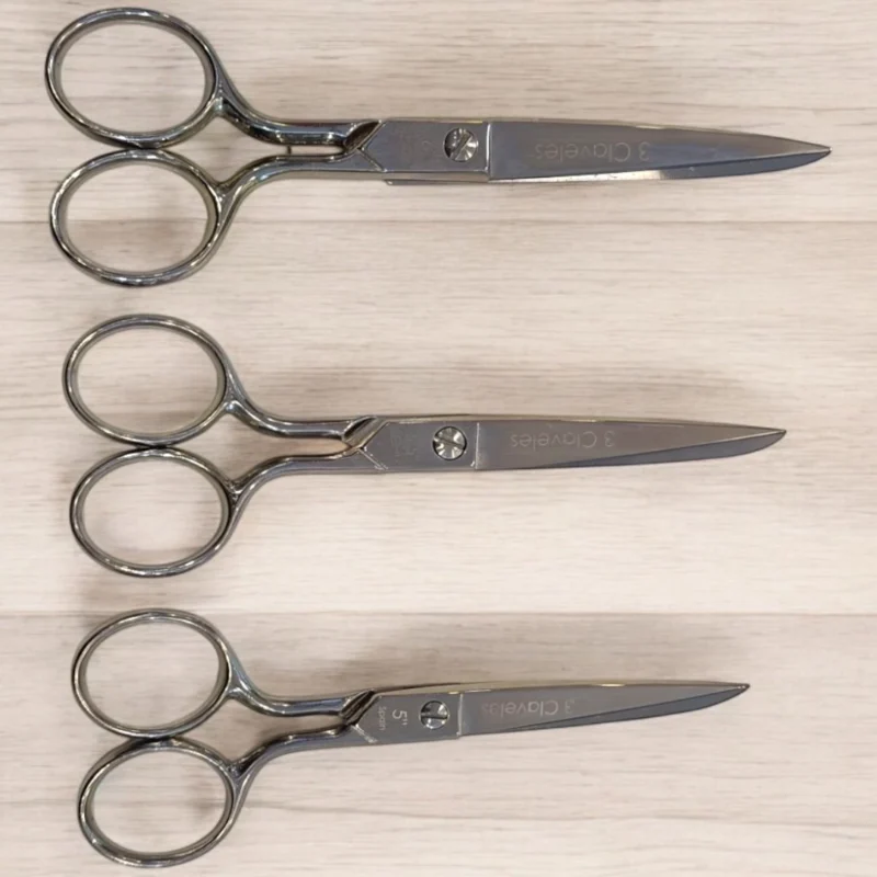 Tres 3 Claveles tijera costura 6" (ref. 00005) están perfectamente alineados horizontalmente sobre una superficie de madera clara, los mangos a la izquierda y las hojas apuntando a la derecha.